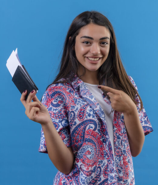 young beautiful woman tourist holding passport with tickets looking at camera pointing with index finger to them smiling cheerfully standing over isolated blue background